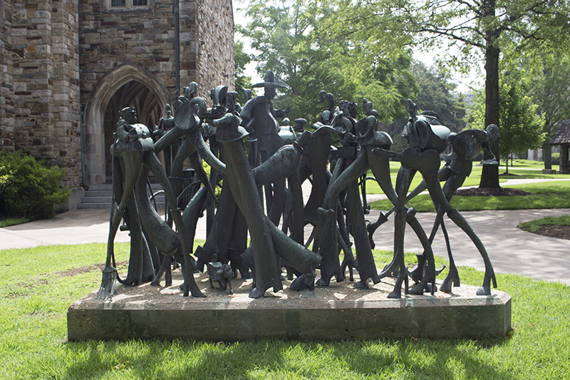 A sculpture consisting of a group of caricatures of people standing and walking as if in a crowd. The figures have very long legs in proportion to their bodies. They wear various articles of clothing including hats, boots, jackets, and pants. They also hold various objects such as books, bags, bicycles, a broom, and a guitar case
