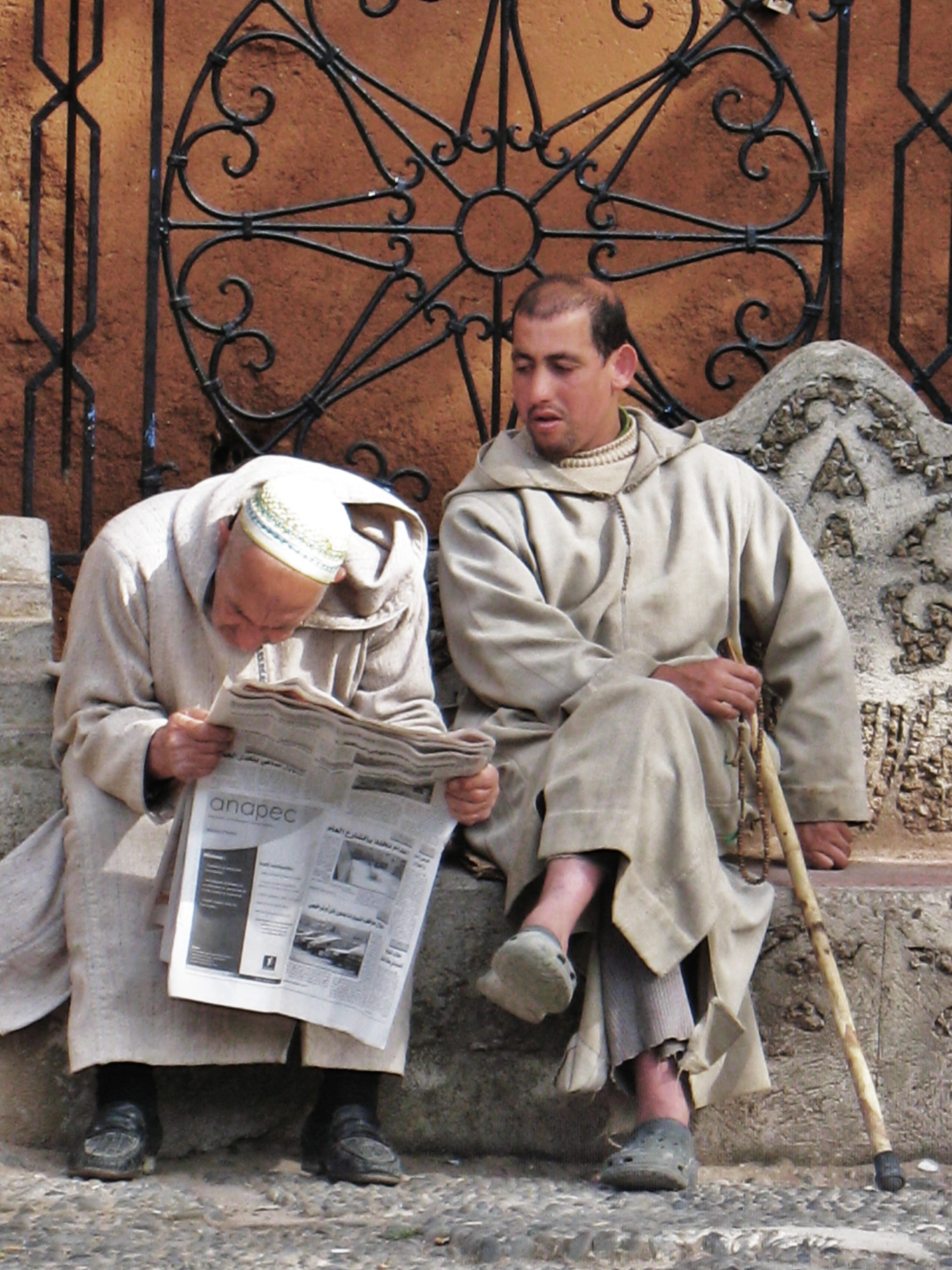 An old man looks closely at the newspaper while his friend tries to read it. 