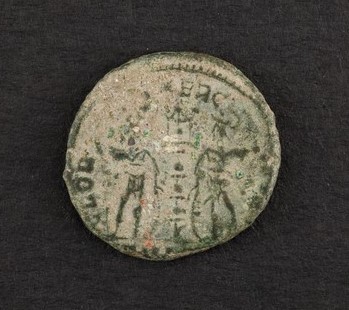 Bronze coin from the Cornell Coin Collection that depicts two soldiers standing between a battle standard. The soldiers look idle, like they are preparing for battle, or a battle will begin soon.