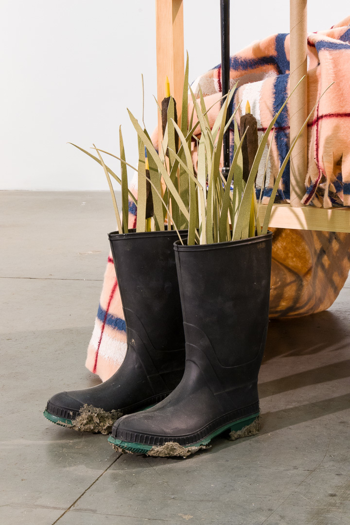 Long grass and catttail plants made of paper stick out of a pair of black rubber boots with dried mud on the bottoms. 