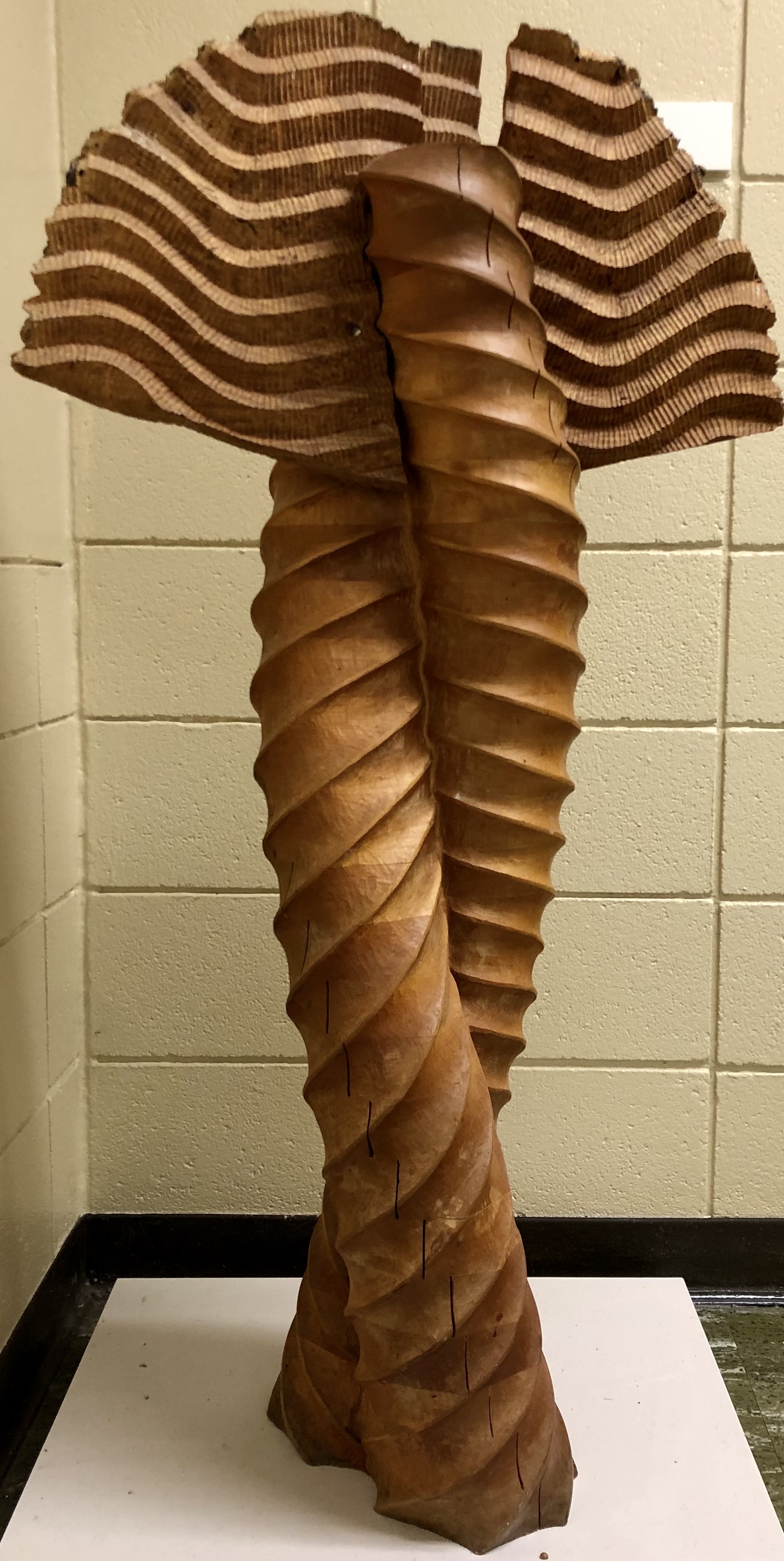 This is a wooden sculpture of two tubular spiraling vertical forms with carved wings attached at the top. 