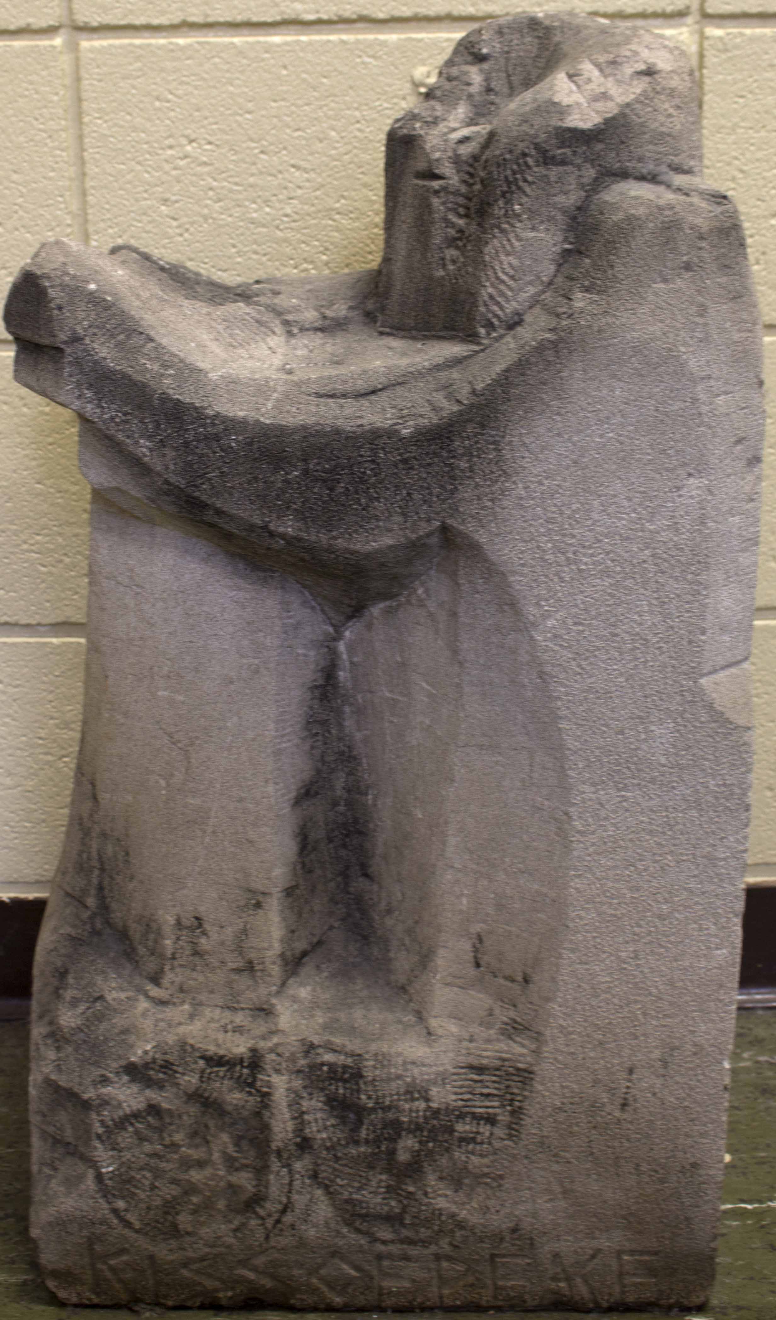 Carved concrete sculpture of a figure with arms extended to the left. At the bottom of the sculpture, it has "KISS OF PEACE" carved. 