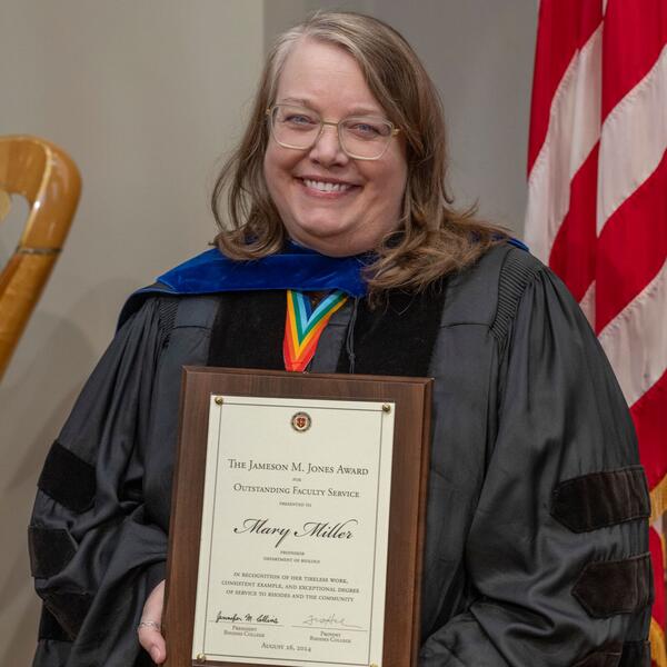a blonde woman in academic robes holds an award plaque
